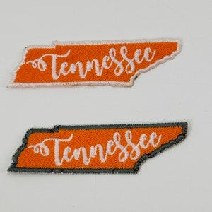 May include: Two orange felt patches shaped like the state of Tennessee with the word "Tennessee" embroidered in white cursive lettering. The top patch has a white outline and the bottom patch has a gray outline.