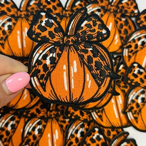 May include: Embroidered orange pumpkin patch with a black and leopard print bow. The pumpkin has white vertical lines and a black outline. A hand with a pink nail is holding one of the patches. The patches are for fall and Halloween.