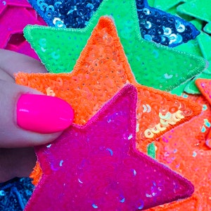 May include: Close-up of colorful sequined star-shaped patches in shades of pink, orange, green, and blue. The stars are embroidered with a contrasting border and are densely covered in sequins, creating a shimmering effect. The image is brightly lit.