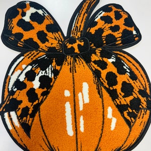 May include: An orange pumpkin-shaped rug with a black outline and a bow featuring a leopard print pattern. The pumpkin has white accents and a textured appearance, perfect for fall decor.