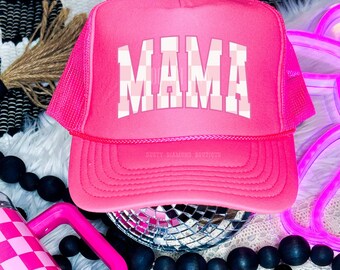 May include: A bright pink trucker hat with a mesh back and a curved brim. The word "MAMA" is printed across the front in a checkered pattern. The text "DTF TRANSFER" is at the top. The hat is from "Dusty Diamond Boutique".