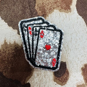 May include: A glittery patch featuring three playing cards, each displaying the Ace of Hearts. The cards have a silver glitter background with black borders and red heart symbols. The patch is likely for clothing or accessories.
