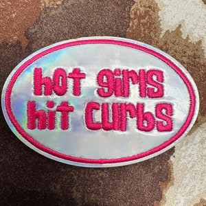 May include: Oval-shaped patch with the text "hot girls hit curbs" in pink, embroidered on a holographic, iridescent background. The patch is outlined in pink and has a textured appearance.