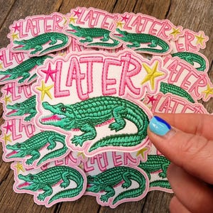 May include: Embroidered patches featuring a green alligator with an open mouth, surrounded by the word "LATER" in pink, with pink stars and yellow stars. The patches have a white background and a pink border.