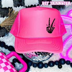 May include: Pink trucker hat with a black skeleton hand making the peace sign. The hat has a mesh back and the text "Dusty Diamond Boutique" is printed on the back.