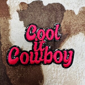 May include: Embroidered patch with the phrase "Cool it Cowboy" in red script lettering outlined in black. The lettering has a glittery silver border. The patch is on a patterned background of brown, tan, and cream.