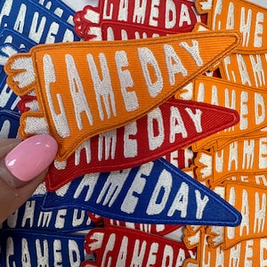May include: A pile of colorful felt pennants with the text "Game Day" in white embroidery. The pennants are in orange, red, and blue.