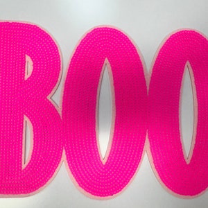 May include: A bright pink, textured "BOO" sign, perfect for Halloween decorations. The letters are in a bold, rounded font, with a dotted texture. The sign is set against a white background.