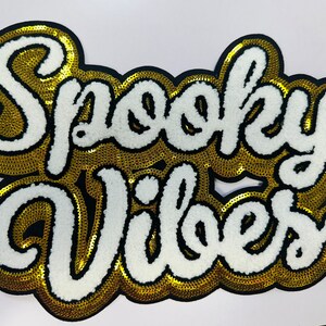 May include: A patch with the words "Spooky Vibes" in white, outlined in gold sequins, and a black border. The patch has a puffy, textured appearance, and is likely for clothing or accessories.
