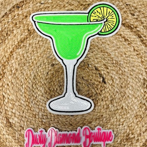 May include: Green margarita glass with a lime wedge on the rim. The glass is made of sequins and has a white base. The image is on a brown woven background with the text "Dusty Diamond Boutique" in pink.