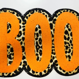 May include: A Halloween-themed patch with the word "BOO" in orange, textured letters. The letters are set against a leopard print background and outlined with a black border. The patch is designed for decorative use.