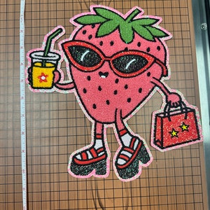 May include: A whimsical, glittery strawberry patch wearing sunglasses and platform sandals, holding a drink and shopping bag. The strawberry is pink with a green stem and is outlined in a sparkly, iridescent material. The shopping bag has two yellow stars.