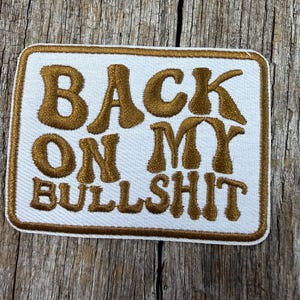 May include: A rectangular patch with the phrase "BACK ON MY BULLSHIT" embroidered in brown on a white background. The patch has a brown border and is displayed on a wooden surface.