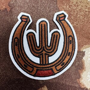 May include: Embroidered patch featuring a brown horseshoe design with a cactus in the center. The patch has a white border and is stitched with brown and black thread. The cactus and horseshoe are detailed with a red accent at the bottom.