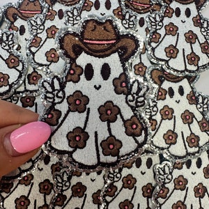 May include: Embroidered patches featuring a cartoon ghost wearing a brown cowboy hat with a pink band. The ghost is white with brown flower details and a silver glitter border. The ghost is giving a peace sign.