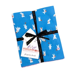 Puede incluir: Un paquete de cuadrados de tela con un fondo azul y un estampado de conejos blancos. El paquete está atado con una cinta negra y una etiqueta que dice "PERSIMMON" y "fat quarter bundle".