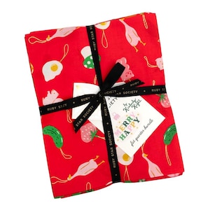 May include: A red fat quarter bundle tied with a black ribbon. The fabric features a whimsical pattern of fried eggs, strawberries, and other food items. A tag reads "Merry Happy" and "fat quarter bundle".