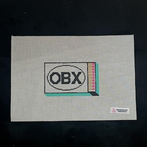 Puede incluir: Un lienzo de punto de aguja con las letras "OBX" dentro de un &oacute;valo negro, dentro de un rect&aacute;ngulo con bordes negros. El rect&aacute;ngulo tiene una base turquesa y un lado a rayas con rosa, amarillo y blanco. El lienzo es gris claro.