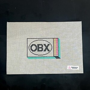 Puede incluir: Un lienzo de punto de aguja con un patr&oacute;n de cuadr&iacute;cula. El dise&ntilde;o presenta las letras "OBX" dentro de un &oacute;valo negro, con un acento rectangular colorido. Una peque&ntilde;a etiqueta en la esquina inferior derecha dice "MOUNTAIN FOX NEEDLEPOINT".
