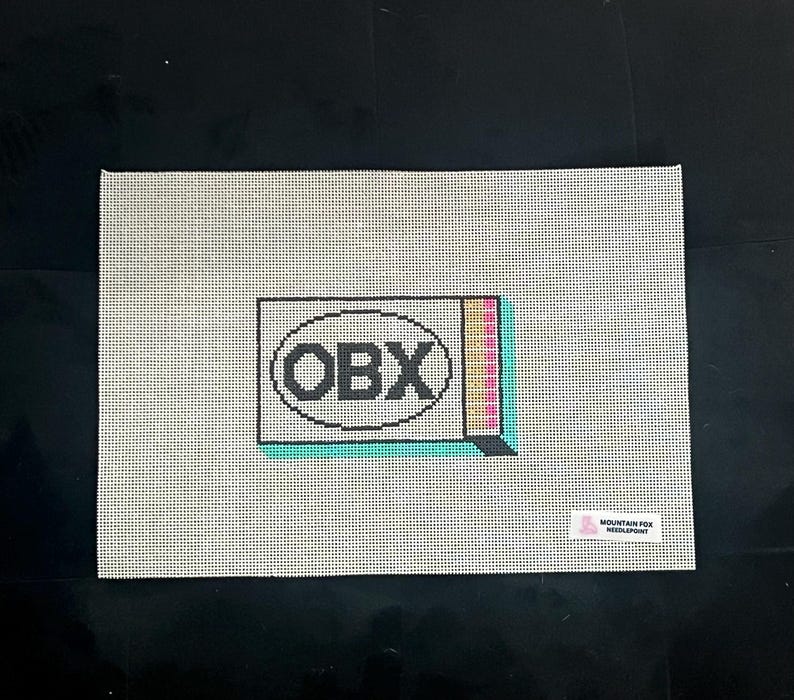 Puede incluir: Un lienzo de punto de aguja con un patr&oacute;n de cuadr&iacute;cula. El dise&ntilde;o presenta las letras "OBX" dentro de un &oacute;valo negro, con un acento rectangular colorido. Una peque&ntilde;a etiqueta en la esquina inferior derecha dice "MOUNTAIN FOX NEEDLEPOINT".