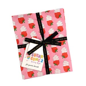 Könnte beinhalten: Ein Stoffpaket, das mit einem schwarzen Band verschnürt ist. Der Stoff hat einen rosa Hintergrund mit einem Muster aus Erdbeeren und Schlagsahne. Ein Etikett trägt die Aufschrift "SUGAR CONE 2" und "fat quarter bundle".