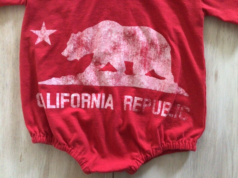 May include: Red baby onesie with a white graphic of the California Republic bear and star. The text "CALIFORNIA REPUBLIC" is printed below the bear.