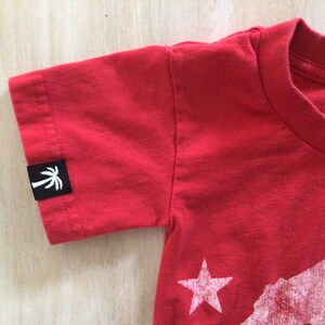 May include: Red short-sleeved t-shirt with a white palm tree logo on the left sleeve and a white graphic of a bear and a star on the front.