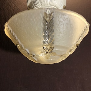 Puede incluir: Una lámpara de techo blanca con una pantalla de vidrio esmerilado que presenta un patrón de hojas decorativas. La pantalla está unida a una base de metal blanco con un borde decorativo.