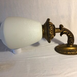 May include: A vintage wall sconce with a white frosted glass shade and a gold-toned metal base. The base features intricate scrollwork and a decorative design.