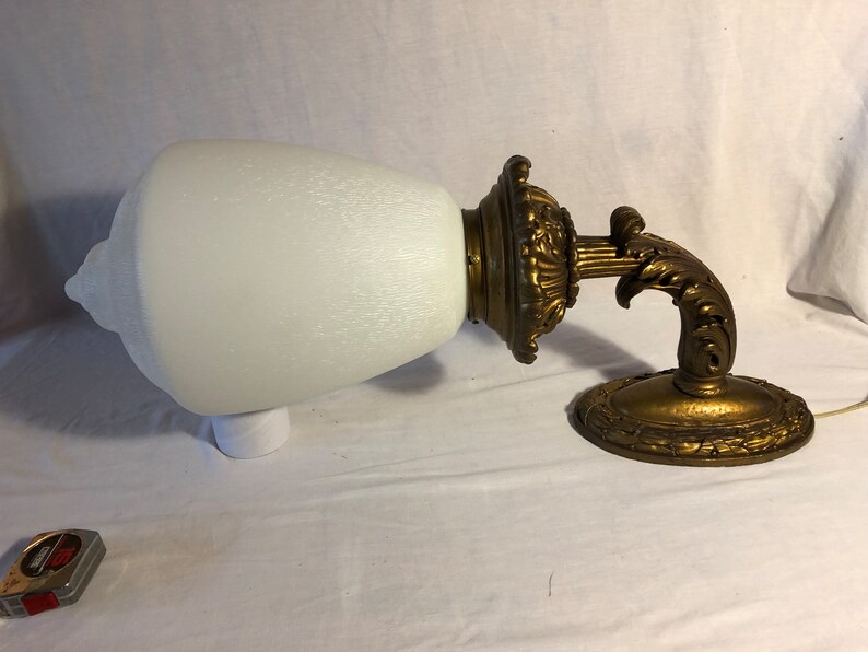 May include: A vintage wall sconce with a white frosted glass shade and a gold-toned metal base. The base features intricate scrollwork and a decorative design.
