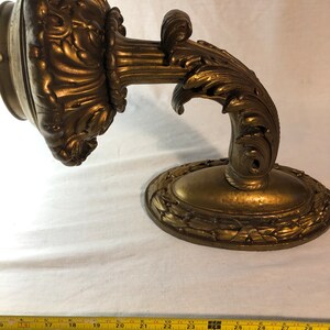 May include: A gold-toned metal wall sconce with ornate scrollwork and a decorative oval base. The sconce is designed to hold a light bulb or candle.