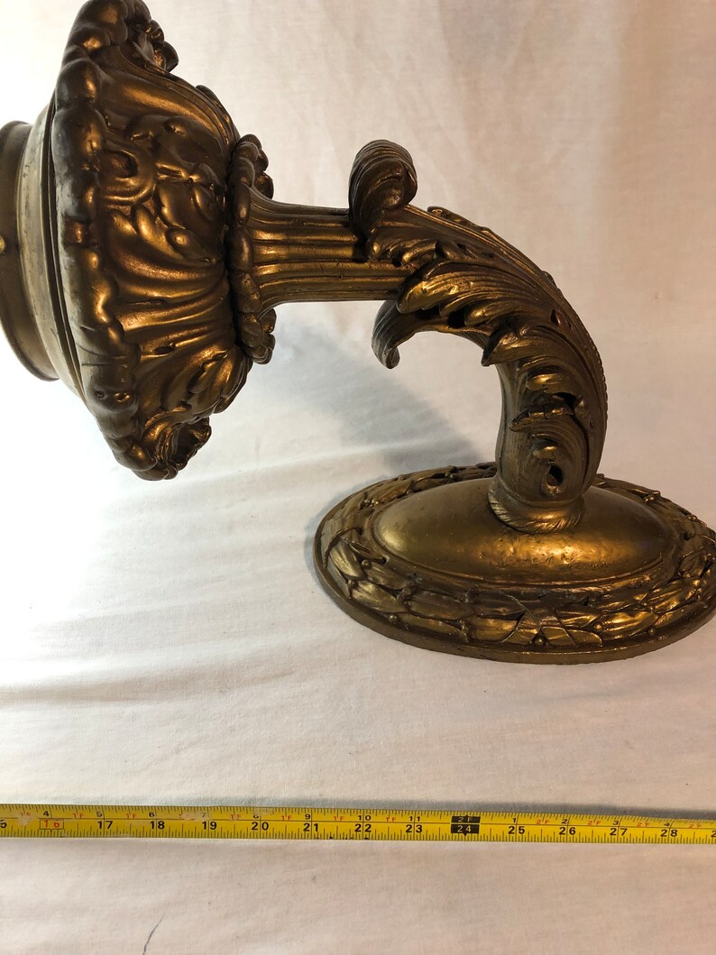 May include: A gold-toned metal wall sconce with ornate scrollwork and a decorative oval base. The sconce is designed to hold a light bulb or candle.