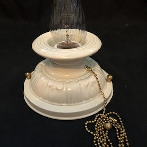 May include: A white ceramic ceiling light fixture with a decorative base and a clear, teardrop-shaped light bulb. A brass pull chain hangs from the fixture. The fixture is on a black background.
