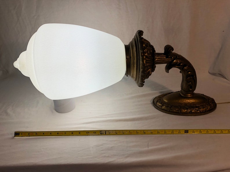 May include: A white frosted glass lampshade with a decorative gold metal base and arm. The lampshade is shaped like a teardrop and the base has intricate details.