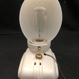 May include: A vintage white ceramic lamp with a curved shade and a visible lightbulb. The lamp has a brass pull chain and a decorative design on the base. The lamp is designed to sit on a table or desk.