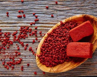 Peut inclure: Un bol en bois rempli de graines d'annatto rouges et de deux blocs d'annatto rouges. Le bol est sur une surface en bois.