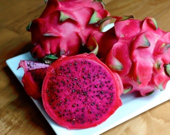 May include: Three dragon fruit, one cut in half, on a white plate. The fruit is pink with green accents and black seeds.