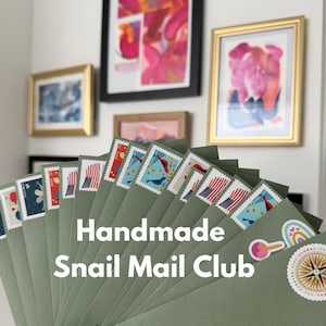 K&ouml;nnte beinhalten: Ein F&auml;cher aus gr&uuml;nen Umschl&auml;gen mit dekorativen Briefmarken, mit dem Text "Handmade Snail Mail Club". Die Briefmarken zeigen verschiedene Designs, darunter Flaggen und V&ouml;gel. Gerahmte Kunstwerke h&auml;ngen im Hintergrund.