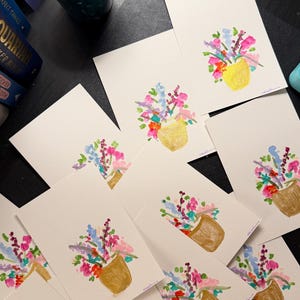 K&ouml;nnte beinhalten: Eine Sammlung von Aquarell-Gru&szlig;karten mit einem Blumenstrau&szlig; in einer goldenen Vase. Die Blumen sind in Rosa-, Blau-, Lila- und Oranget&ouml;nen gemalt. Die Karten sind auf einer dunklen Oberfl&auml;che angeordnet, mit einer Tasse und anderen Objekten im Hintergrund.