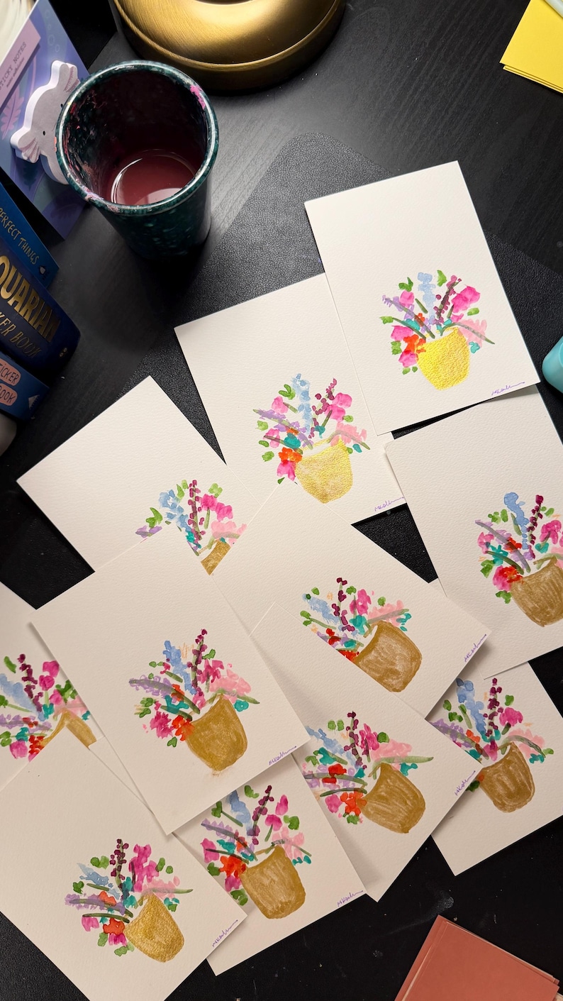 K&ouml;nnte beinhalten: Eine Sammlung von Aquarell-Gru&szlig;karten mit einem Blumenstrau&szlig; in einer goldenen Vase. Die Blumen sind in Rosa-, Blau-, Lila- und Oranget&ouml;nen gemalt. Die Karten sind auf einer dunklen Oberfl&auml;che angeordnet, mit einer Tasse und anderen Objekten im Hintergrund.