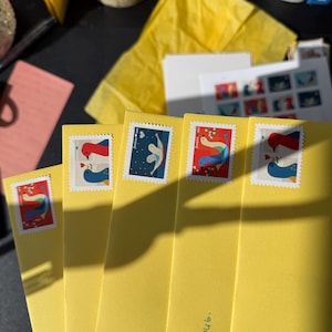 K&ouml;nnte beinhalten: Eine Reihe gelber Umschl&auml;ge mit dekorativen Briefmarken, die Vogelillustrationen zeigen. Die Briefmarken zeigen verschiedene Designs mit roten, blauen und wei&szlig;en V&ouml;geln, einige mit Herzmotiven. Zus&auml;tzliche Schreibwarenartikel sind im Hintergrund zu sehen.