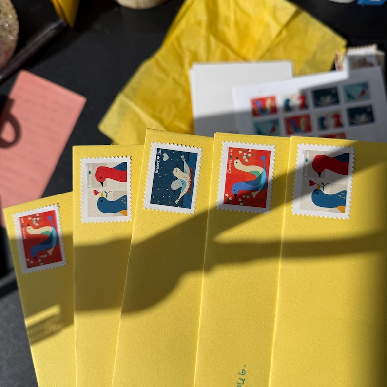 K&ouml;nnte beinhalten: Eine Reihe gelber Umschl&auml;ge mit dekorativen Briefmarken, die Vogelillustrationen zeigen. Die Briefmarken zeigen verschiedene Designs mit roten, blauen und wei&szlig;en V&ouml;geln, einige mit Herzmotiven. Zus&auml;tzliche Schreibwarenartikel sind im Hintergrund zu sehen.