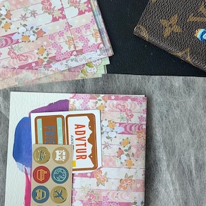 K&ouml;nnte beinhalten: Eine Sammlung von Schreibwaren, darunter Papier mit floralem Muster, Aufkleber und Stifte. Das Papier hat ein rosa-wei&szlig;es Blumenmuster. Eine braune, gemusterte Geldb&ouml;rse ist ebenfalls zu sehen.