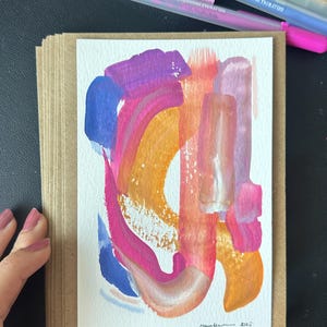 K&ouml;nnte beinhalten: Abstrakte Kunst-Gru&szlig;karte mit farbenfrohem Design. Das Kunstwerk zeigt kr&auml;ftige Pinselstriche in Rosa-, Orange-, Blau- und Lilat&ouml;nen. Die Karte wird in einem Stapel aus Kraftpapierkarten gehalten. Gelstifte und ein Bleistift befinden sich im Hintergrund.