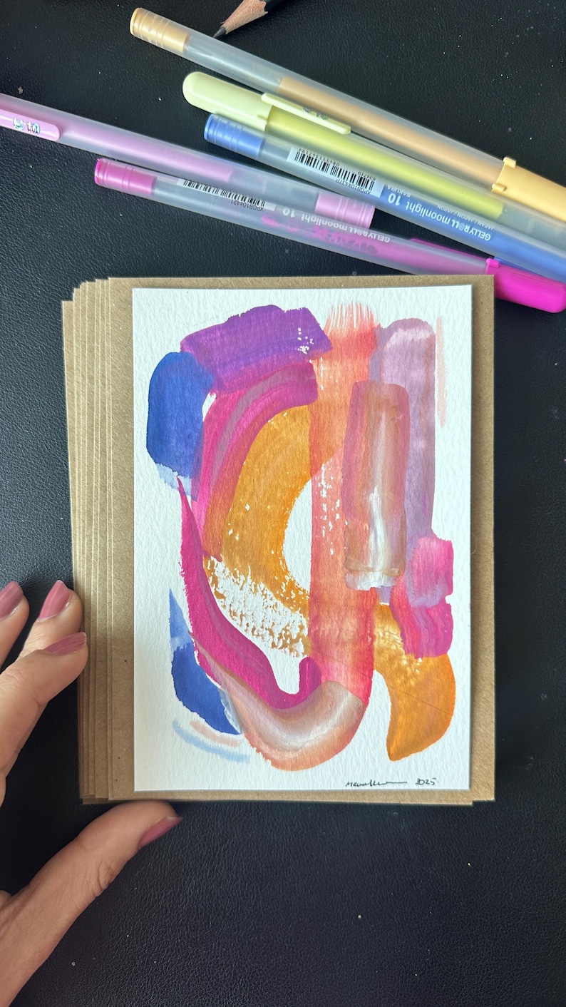 K&ouml;nnte beinhalten: Abstrakte Kunst-Gru&szlig;karte mit farbenfrohem Design. Das Kunstwerk zeigt kr&auml;ftige Pinselstriche in Rosa-, Orange-, Blau- und Lilat&ouml;nen. Die Karte wird in einem Stapel aus Kraftpapierkarten gehalten. Gelstifte und ein Bleistift befinden sich im Hintergrund.