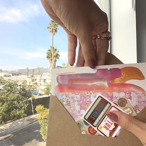 K&ouml;nnte beinhalten: Eine Hand h&auml;lt eine Gru&szlig;karte mit abstrakten Pinselstrichen in Rosa, Orange und Lila. Die Karte befindet sich in einem braunen Umschlag mit Aufklebern mit der Aufschrift "FRIENDS" und "NATURE". Der Hintergrund zeigt eine sonnige Stadtlandschaft mit Palmen.
