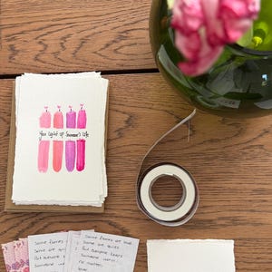 K&ouml;nnte beinhalten: Handgemachte Gru&szlig;karten mit Aquarell-Kerzenillustrationen in Rosa- und Lilat&ouml;nen. Der Text "You light up Someone's Life" ist auf den Karten geschrieben. Eine Rolle doppelseitiges Klebeband und eine Vase mit rosa Tulpen sind ebenfalls zu sehen.