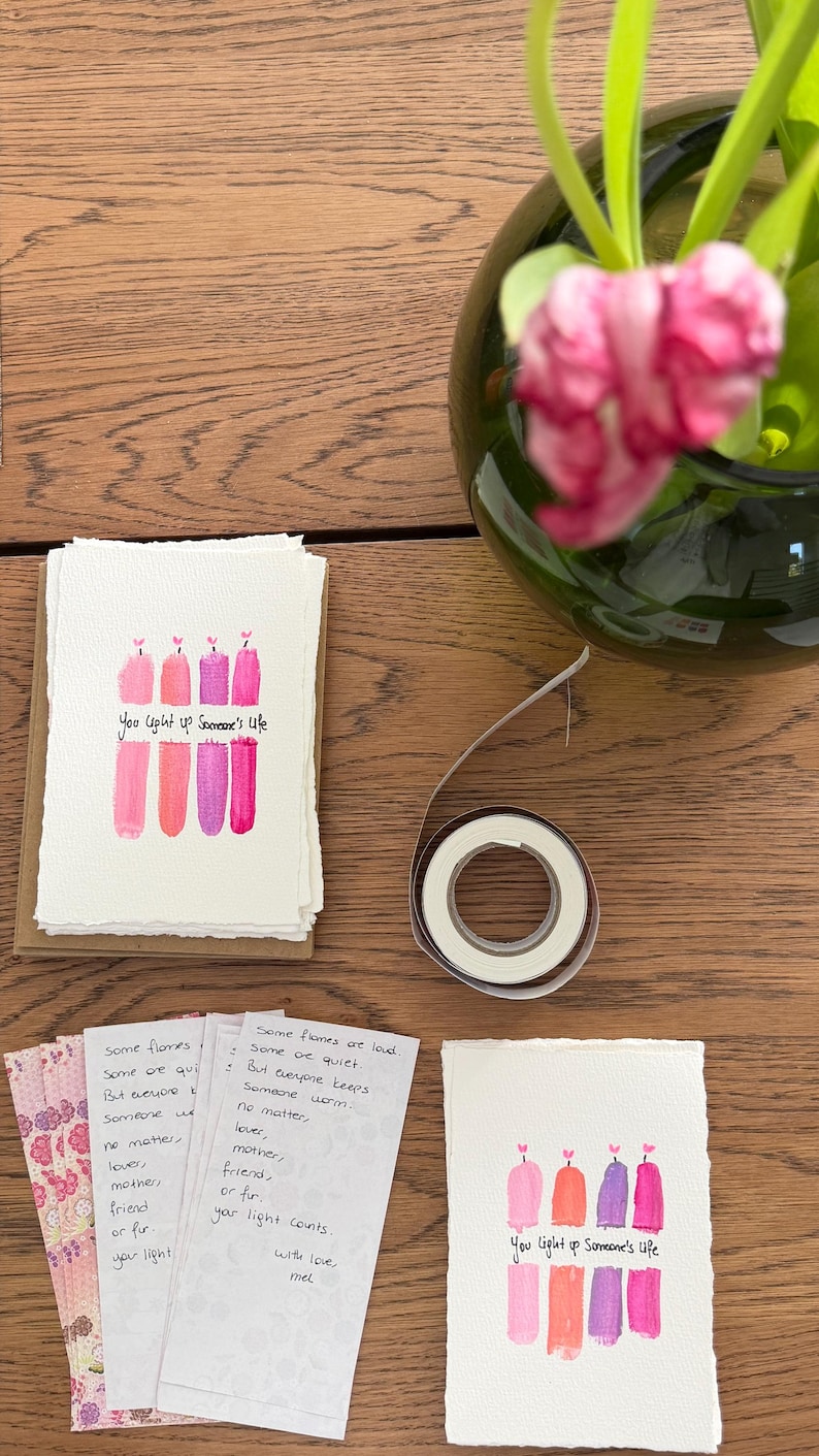 K&ouml;nnte beinhalten: Handgemachte Gru&szlig;karten mit Aquarell-Kerzenillustrationen in Rosa- und Lilat&ouml;nen. Der Text "You light up Someone's Life" ist auf den Karten geschrieben. Eine Rolle doppelseitiges Klebeband und eine Vase mit rosa Tulpen sind ebenfalls zu sehen.