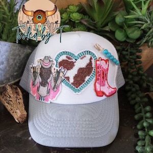Op de afbeelding: Witte truckercap met een grijze klep. De pet heeft drie geborduurde patches: een cowgirl met een roze shirt, een bruin en wit koeienhart en een roze cowboylaars. De pet heeft ook een turquoise kralenketting.