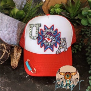 May include: A red and white trucker hat with a white mesh back. The hat has a large, sparkly, blue, red, and white "USA" design with a turquoise and white beaded safety pin attached to the brim.